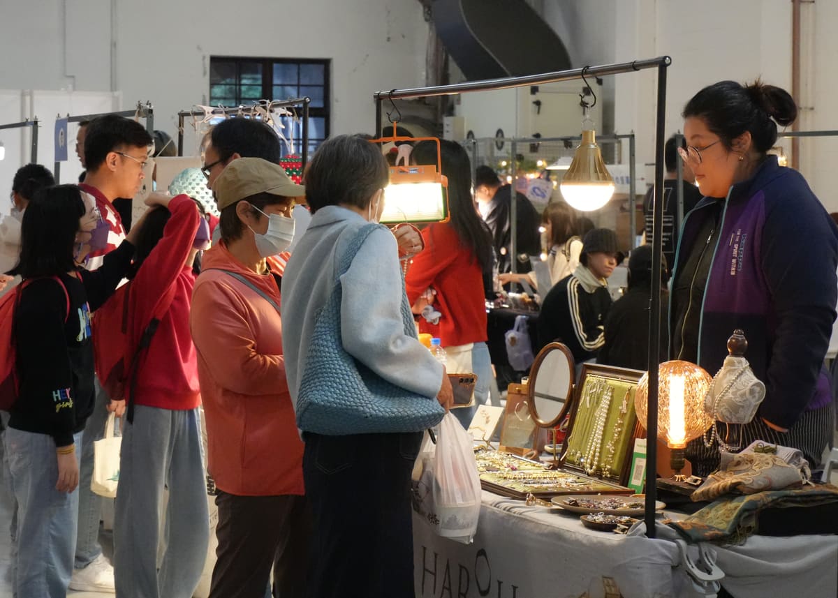 活動花絮：民眾排隊在飾品攤前選購，攤主在燈泡與珠鍊首飾展示架後方向顧客介紹