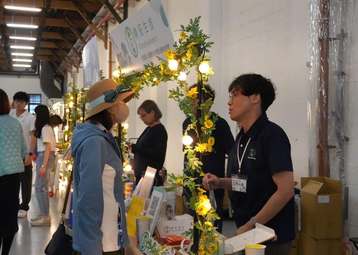 活動花絮：戴帽子與口罩的民眾在精拓生技攤位前，與穿著深藍色工作人員背心的人員交談，攤位以黃色花藤與小燈泡裝飾