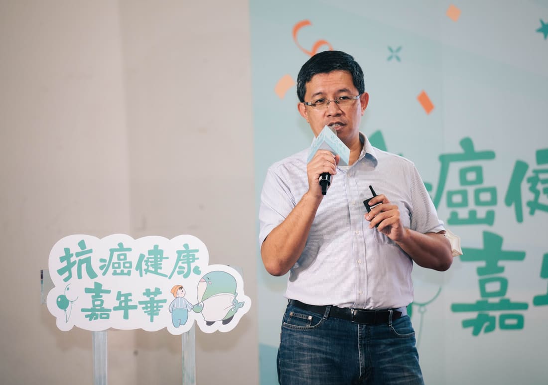 照片：講者手持麥克風在活動舞台上分享，左下方立牌寫著「抗癌健康嘉年華」並印有鯨魚吉祥物